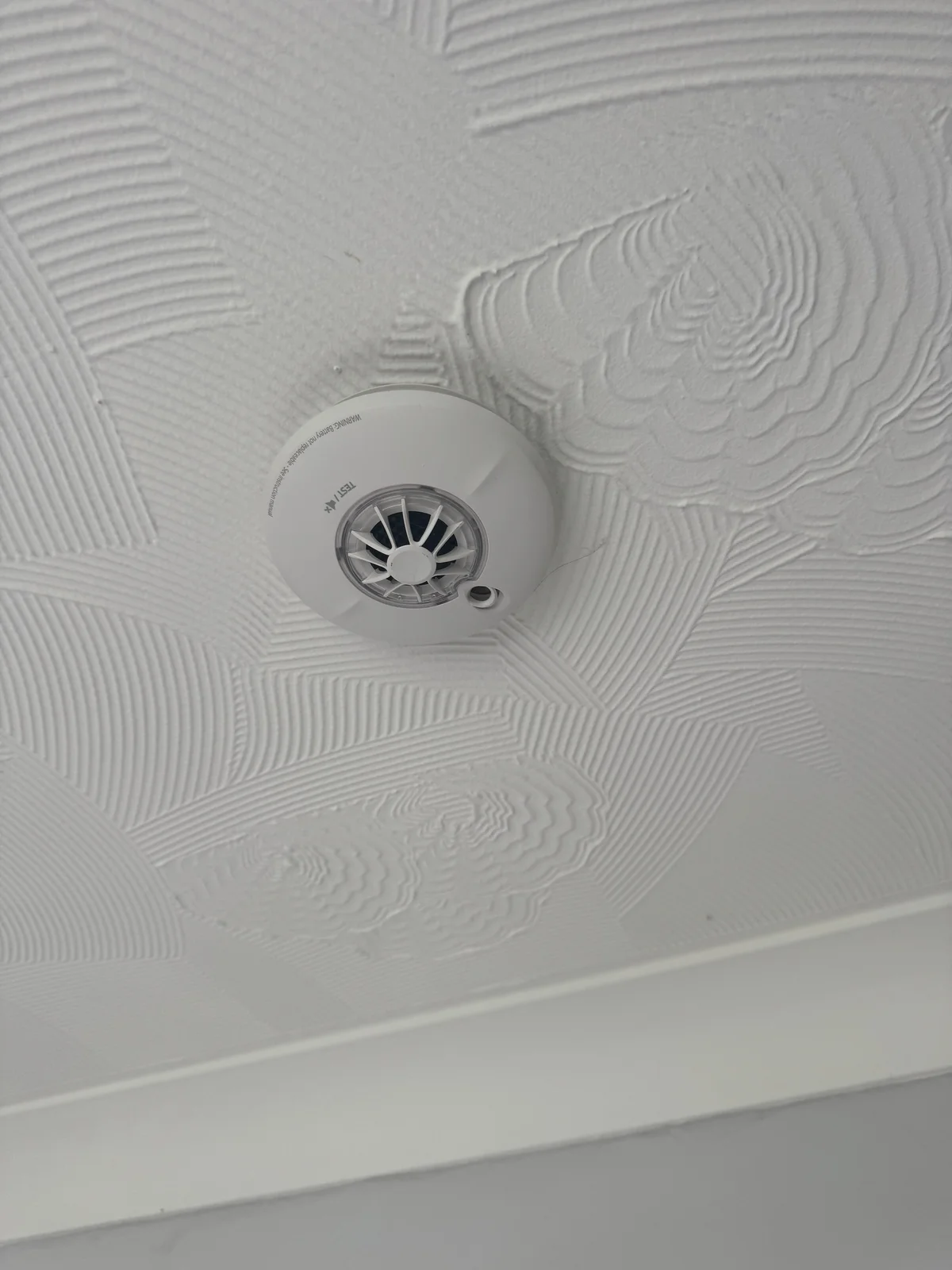 A white ceiling with combed and swirled surface texture around a ceiling-mounted alarm.
