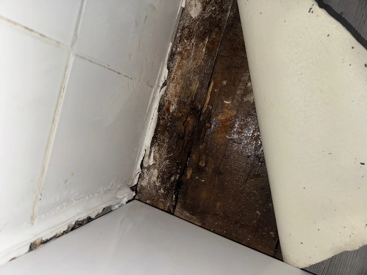 A turned-back floor covering exposes darkened timber boards beside a tiled bathroom wall and a white fitting.